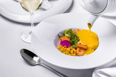 Karidesli çorba, mısır, çiçek yaprakları, taze filizler, çatal bıçaklı bir tabak yeşil bezelye ve masada bir bardak şarap.