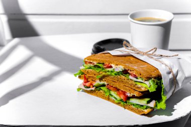 Sandwich in white paper with ham, lettuce, tomatoes, cucumbers and sauce with a paper cup of cappuccino and a black lid standing on a white chair against a background of a white wooden wall