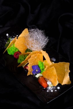 Cips, böğürtlen, böğürtlen, ahududu kremalı şeker ve siyah bir tabakta şeker iplikleri ve koyu bir arka plan. Dikey yönelim