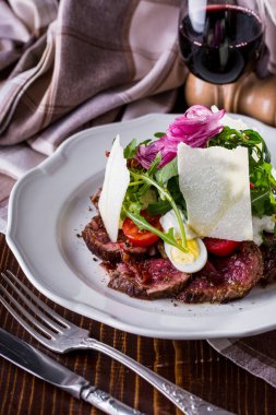 Salad with beef meat, tomatoes, boiled egg, arugula, onion, cheese, salad and sauce on a wooden background with cutlery and a glass of wine