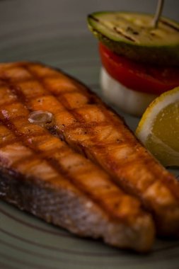 Grilled salmon steak with lemon slice, fried avocado, pominador slice and onion rings on a round plate and wooden table. Vertical orientation 