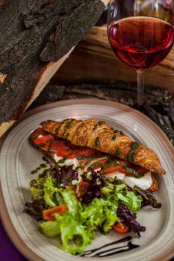 Croissant with mozzarella, lettuce, tomatoes and pesto. Next to it is a mix of green and dark salad with cherry tomatoes and pesto sauce. They lie on a ceramic plate. The plate is on a wooden table, there is wood for the fireplace and a glass of rose
