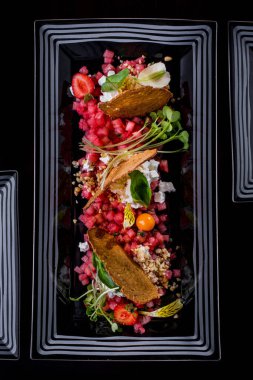 İnce doğranmış karpuz, ekmek kızartması, Philadelphia peyniri, fesleğen yaprağı, mikroyeşil fesleğen, fındık kırıntıları ve çilek. Salata dikdörtgen şeklinde, beyaz desenli siyah bir tabakta. Plaka siyah bir arkaplanda duruyor.