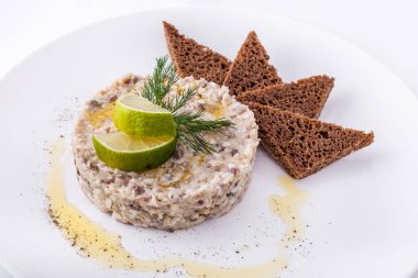 Ringa balığı. Üstte limon kamaları ve dereotundan bir filiz var. Yanında üç tost çavdar ekmeği var. Yemek beyaz bir tabakta, zeytinyağı ve biber tabağa serpilmiş. Plaka beyaz bir arkaplanda duruyor..