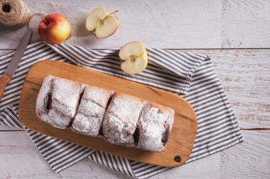 Sweet white bread with powdered sugar, apples lying on a wooden board with a towel and a knife on the table