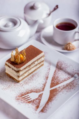 Bir fincan kahve, şeker ve kremayla birlikte beyaz bir tabağa bir dilim tiramisu ve kakao serpiştirilmiş. Dikey yönelim