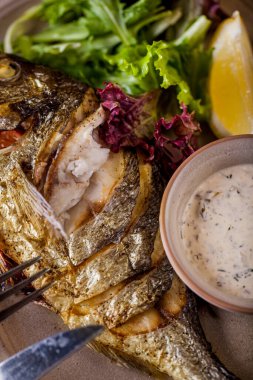 Fried and grilled dorado with white sauce with parsley, a slice of lemon, mung bean salad, arugula on an oval plate with a glass of wine. Vertical orientation 