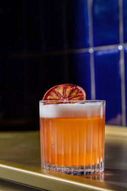 Old fashioned alcoholic cocktail with Sicilian orange chips, single malt whiskey and a large block of ice. Cocktail in a crystal, faceted glass. The glass stands on a golden metal stand, with a blue ceramic tile behind the background.