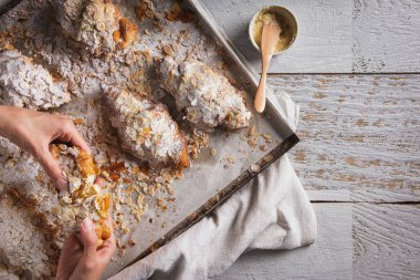 Croissants with condensed milk are spread by hands with powdered sugar and almond slices lying on parchment and a tray with a towel and filling in a bowl.