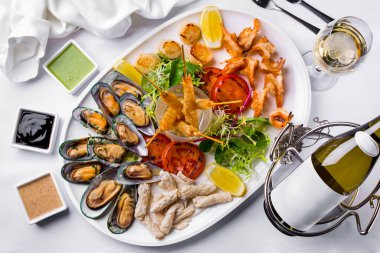 Kızarmış balık, midye, domates, limon, marul parçaları. Genç filizler, fesleğenler, karidesler, ahşap bir tabakta balık fileto, bir şişe şarap ve arka planda üç farklı sos. Yatay yönelim