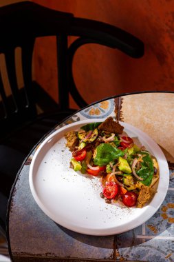 Salad with zucchini, onions, tomatoes, lettuce, croutons, cucumbers and sauce in a plate on the table against a brown wall