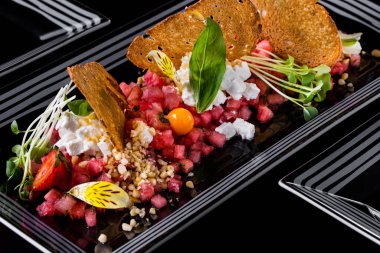 İnce doğranmış karpuz, ekmek kızartması, Philadelphia peyniri, fesleğen yaprağı, mikroyeşil fesleğen, fındık kırıntıları ve çilek. Salata dikdörtgen şeklinde, beyaz desenli siyah bir tabakta. Plaka siyah bir arkaplanda duruyor.