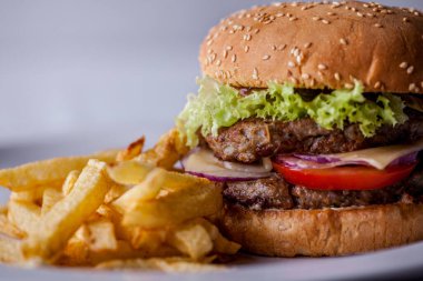 Burger with sesame bun, onion, salad, two meat patties, tomatoes and cheese on a wooden board with fries. Horizontal orientation
