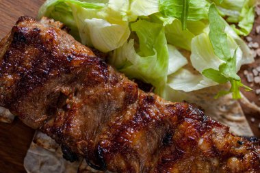 Grilled pork ribs in honey sauce. They lie on pita bread. Nearby is a Peking cabbage salad, a sprig of thyme and a beige ceramic sauce bowl with homemade adjika. Food and sauce stand on a wooden board. The board is on the table, next to a pepper shak