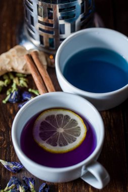 Tayland mavi çayı Chang Shu beyaz seramik bardakta, yanında bir dilim limonlu bir bardak daha var, Chang Shu 'nun içinde mor var. Çanak çömlek tahta bir arka planda duruyor. Yanında Chang Shu 'nun demlediği çay makinası var. Kurutulmuş çiçek.