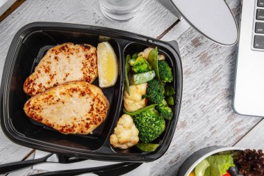 Fried cutlet with lemon, broccoli in a plastic container with a glass of water, and a laptop standing on a white wooden table