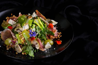Prosciutto salad, grapes, lettuce, arugula, grains, for blue cheese, flower petals in a plate on a dark background