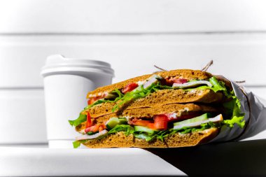 Sandwich in white paper with ham, lettuce, tomatoes, cucumbers and sauce with a paper cup of espresso with a lid standing on a white chair against the background of a white wooden wall