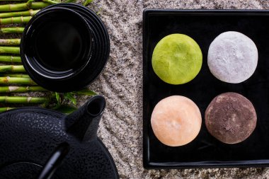 Siyah bir tabakta dört mochi ve bambu kaplı bir çay seti. Yatay yönelim