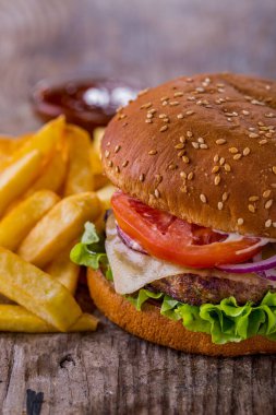 Burger with sesame bun, cutlet, tomatoes, onions, lettuce, cheese and mayonnaise with fries and sauce on a wooden table. Vertical orientation