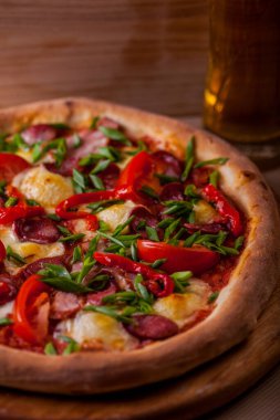 Pizza with cherry tomatoes, ham, sausages, green onions and cheese on a wooden board with a glass of beer on the table