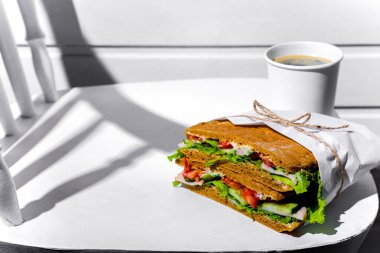 Sandwich in white paper with ham, lettuce, tomatoes, cucumbers and sauce with cappuccino with a paper cup standing on a white chair against the background of a white wooden wall
