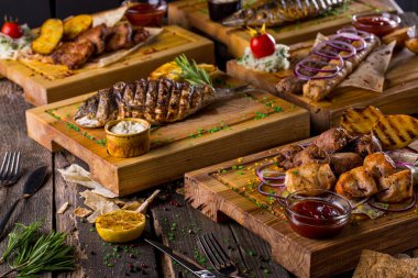 Grilled dorado with, a slice of lemon, rosemary, pieces of pita bread, a glass of juice, salt, pepper on a wooden board, fried potatoes, pieces of pork and chicken skewers, kebab with white sauce, ketchup with cutlery on a wooden table. Horizontal or