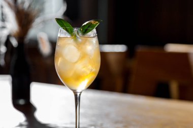 A cocktail with ice, syrup and basil in a glass that stands on the edge of a wooden table surrounded by a restaurant