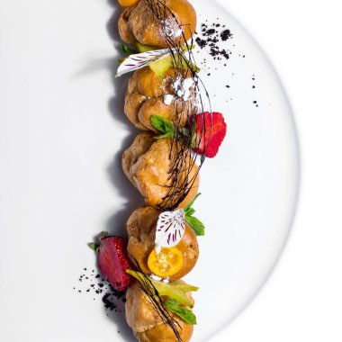 Kremalı profiteroller, kumkat, çilek, nane, kumkat, çiçek yaprakları ve beyaz bir tabakta pudra şekeri.