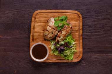 Üstünde nane yaprakları olan ızgara hindi filetosu. Yanında salata, kırmızı biber halkaları ve erik soslu sos kabı var. Yiyecekler ahşap, kare bir tahtanın üzerinde. Tahta kahverengi, ahşap bir arka planda duruyor..