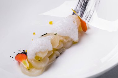 Kafa derisi, ince dilimlenmiş, kaydırağa istiflenmiş, üzeri narenciye köpüğü ve yenilebilir yapraklarla kaplı. Yemek derin beyaz bir tabakta. Tabak beyaz bir masa örtüsünün üzerinde, yanında çatal bıçak takımı ve bez peçeteler var, ve bir bardak gül şarabı var..