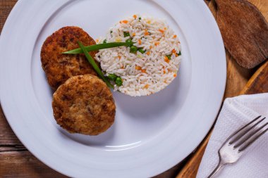 Two veal cutlets with green onions, next to rice with green peas, carrots and zucchini. The food is on a white plate. The plate stands on a wooden background, a fork and knife, a spatula for a frying pan lie next to it.