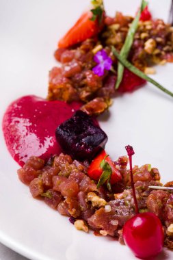 Çilek reçelli ton balığı tartartarı, çilek dilimleri, kokteyl kirazı, fıstık, yanmış şeker cipsi. Yeşil soğan ve karamelli pancar dilimleri. Yemek beyaz, derin bir tabakta. Tabak beyaz bir masa örtüsünün üzerinde, yanında çatal bıçak takımı var, beyaz bir c harfi.