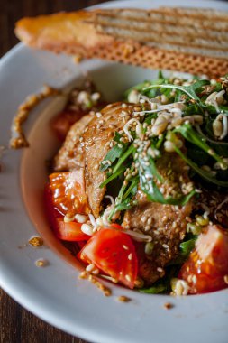 Salad with ham meat, sesame seeds, young shoots, arugula, sauce, tomatoes and lettuce, bread in a plate on a wooden background