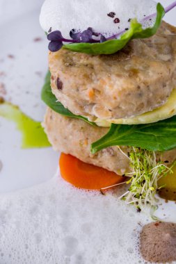 Steamed fish cakes with spinach leaves, knee cucumber, carrots and citrus foam. The fish is on a white ceramic plate. The plate is on a stand with a beige tablecloth, next to it is a cloth napkin, a fork with a knife, two glasses of white wine and an