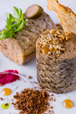 Sturgeon stuffed with bread chips, peanuts, parsley sprig and capers. The fish is on a white ceramic plate. The plate is on a stand with a beige tablecloth, next to it is a cloth napkin, a fork with a knife, two glasses of white wine and another plat