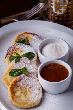 Beyaz bir tabakta pudra şekeri ve nane şekerli Syrniki. Yanında kayısı reçelli bir sos kasesi ve ekşi kremalı bir sos kasesi var. Tabak ahşap bir masanın üzerinde, yanında deniz dikenli çay makinası ve çatal ve bıçak var. 