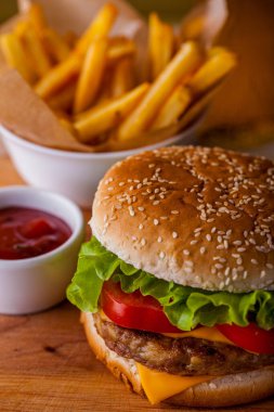 Burger with sesame bun, onion, lettuce, meat cutlet, tomatoes and cheese on a wooden board with sauce and fries. Vertical orientation