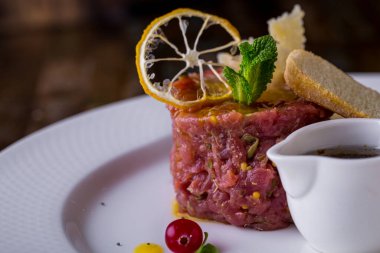 Dijon hardalı ve mango soslu dana tartar. Üstte limon cipsi, nane yaprağı, parmesan dilimi ve beyaz ekmek tostu var. Yakınlarda salamura ve karamelli soğanla dolu bir sos kabı var. Yemekler beyaz tabakta..