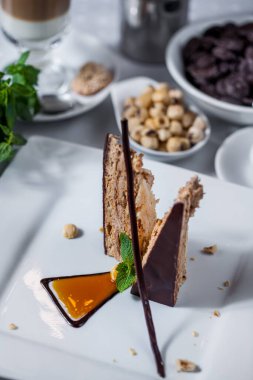 Waffle katmanlı bir dilim pasta çikolata jöleli, naneli, karamelli fındıklı, beyaz tabakta çikolata parçacıklı bir fincan kahve ve krema. Dikey yönelim