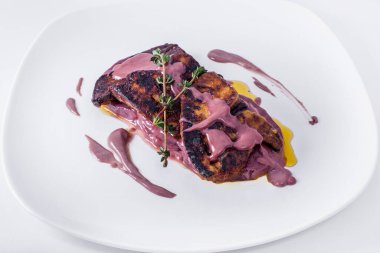 Kızarmış kaz ciğeri ezmesi, vişne soslu ananas ve kekik soslu. Tabağa biraz kiraz sosu bulaşmış. Kaz ciğeri beyaz bir terliğin üzerinde, tabak beyaz bir arka planda duruyor..