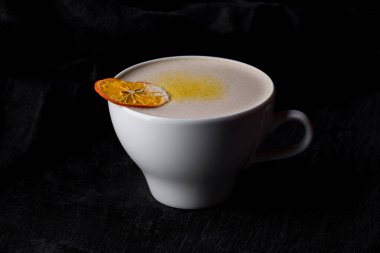 Sütlü kakao, portakal cipsi ve turunçgil soslu. Seramik beyaz bardakta iç. Bardak siyah, ahşap bir zemin üzerinde duruyor, arkasında siyah keten bir kumaş var..