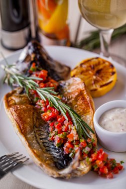 Grilled stuffed mackerel with lemon, rosemary, sauce in a gravy boat on an oval white plate and a glass of white wine on a wooden table. Vertical orientation 