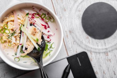 Okroshka with a spoonful of radishes, cucumbers, onions, eggs and ham standing on a table with a tablet