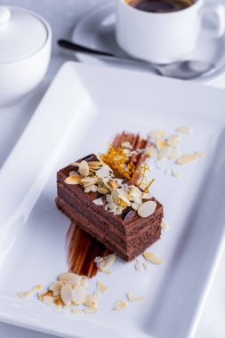 Bir dilim çikolatalı pasta, karamel ve fındık parçalarını beyaz tabakta bir fincan kahveyle birlikte. Dikey yönelim
