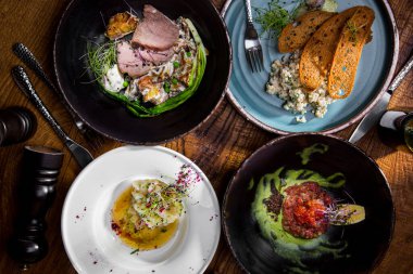 On a wooden table there are cutlery, a pepper shaker, a salt shaker, a plate with minced meat, salmon caviar, sprouts, lemon and sauce in a plate, fried bread, with sprouts, onions, salad, and a boiled egg in the second plate, squid meat, sprouts and