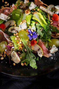 Sproshutto salad, grapes, lettuce, arugula, grains, dor blue cheese, flower petals in a plate on a dark background