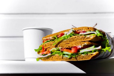 Sandwich in white paper with ham, lettuce, tomatoes, cucumbers and sauce with cappuccino with a paper cup standing on a white chair against the background of a white wooden wall