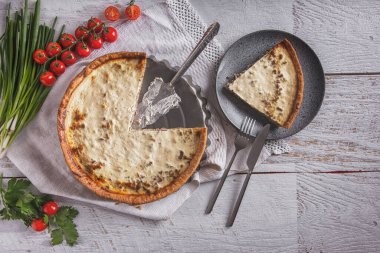 Cheese casserole with tomatoes, onions and parsley on a towel with cherry sprigs and a piece of casserole in a plate lying on the table