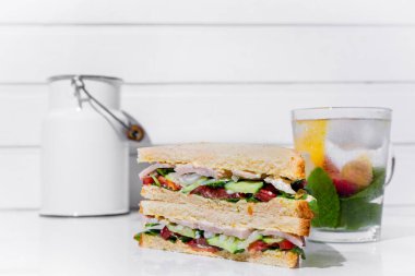Sandwich with ham, onions, cucumbers, tomatoes, sauce and salad and a cooling cocktail with orange, passionfruit, mint and ice in a glass with a white can on a white table on a white wooden wall background